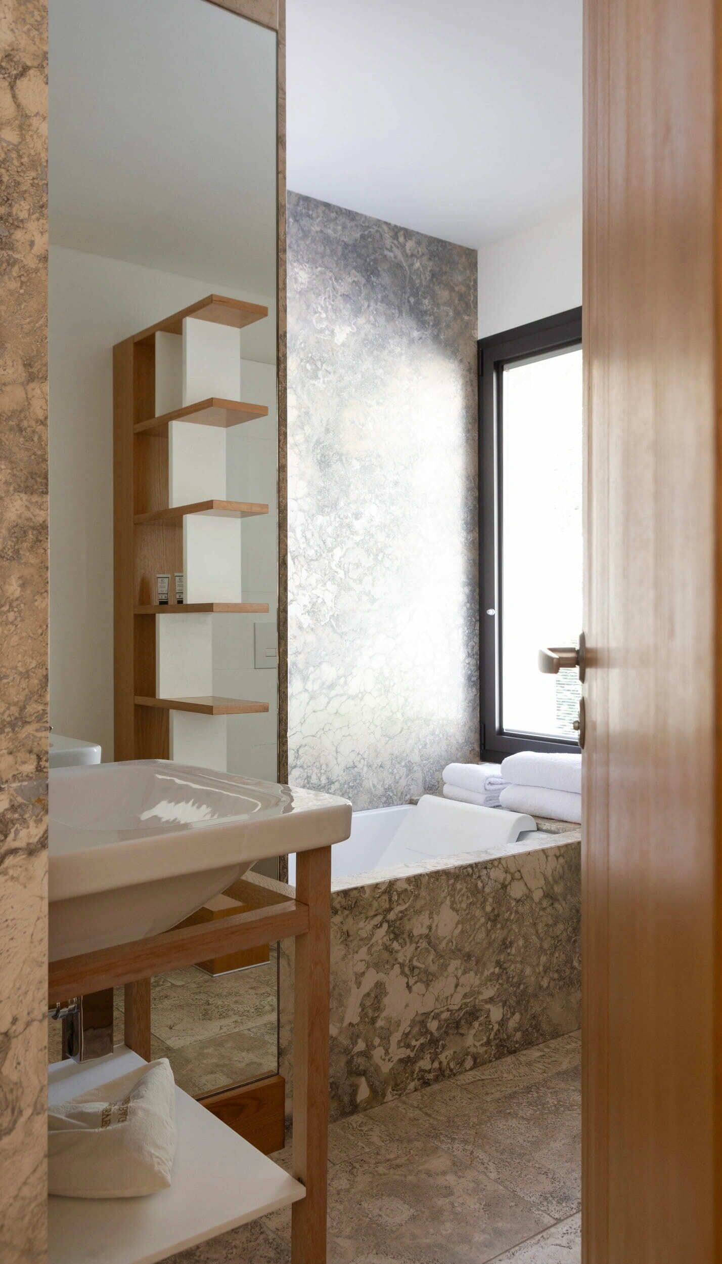 A bathroom with a marble sink, a wooden shelf, a large mirror, and a bathtub with towels.
