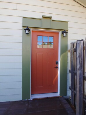 A red door with a small window, flanked by two wall-mounted lanterns, set against a green and white exterior wall. - Comfortable private guest suite (Flagstaff)