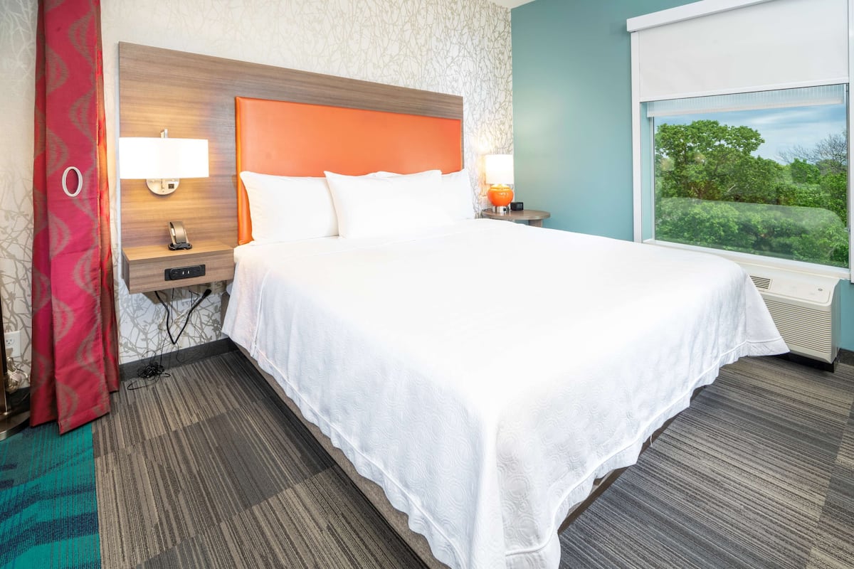 A hotel room with a large bed, two bedside tables with lamps, a window with a view of greenery, and a patterned carpet.