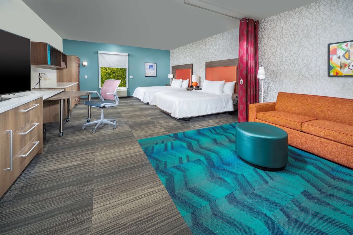 A hotel room with a desk, two beds, a sofa, a TV, and a view of greenery.