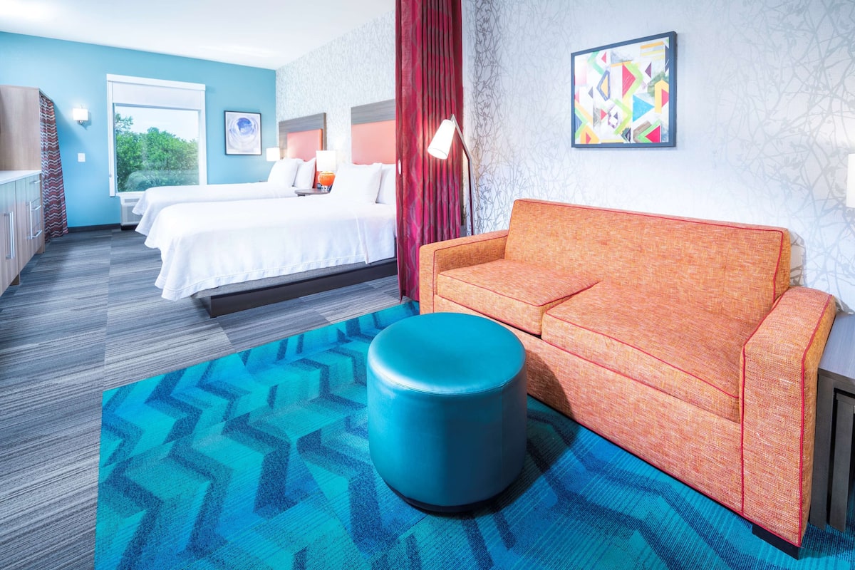 A hotel room with a bed, a sofa, a blue ottoman, and a patterned rug.