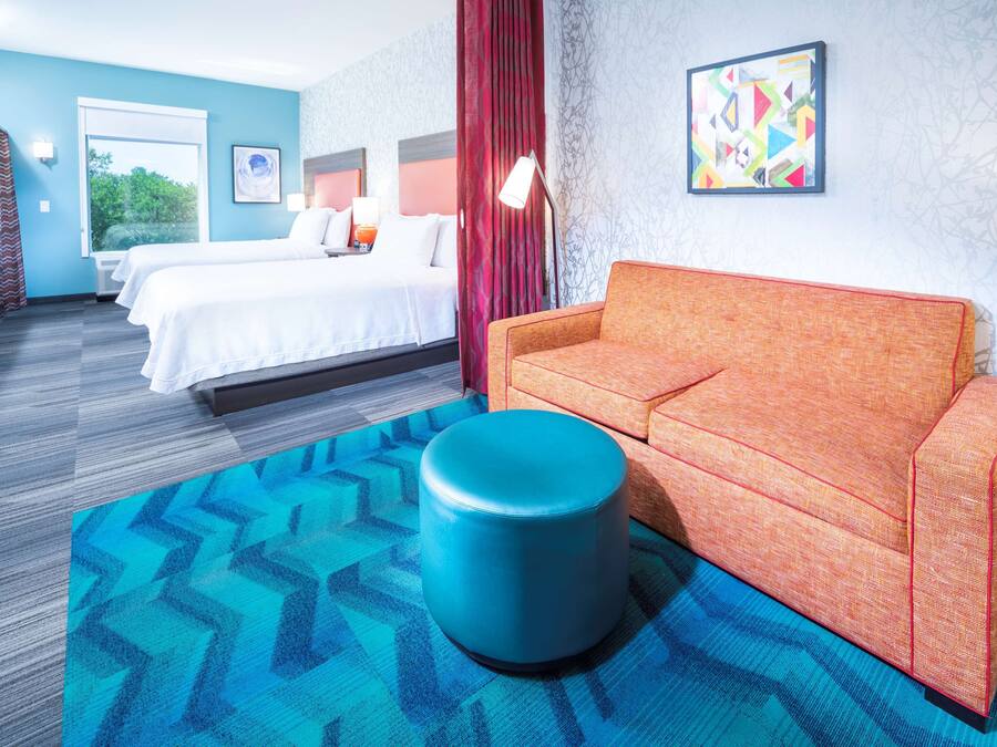 A hotel room with a bed, a sofa, a blue ottoman, and a patterned rug.