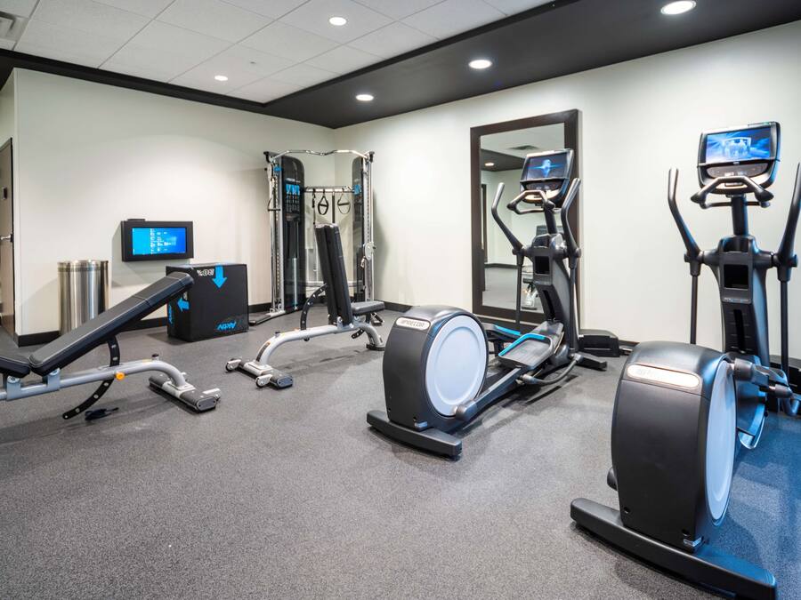 Academia com máquinas de cardio, equipamentos de musculação e uma tela de televisão.