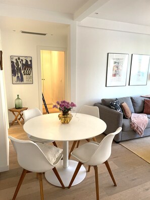 A modern living room with a round white table, white chairs, a grey sofa, and a green vase with purple flowers.
