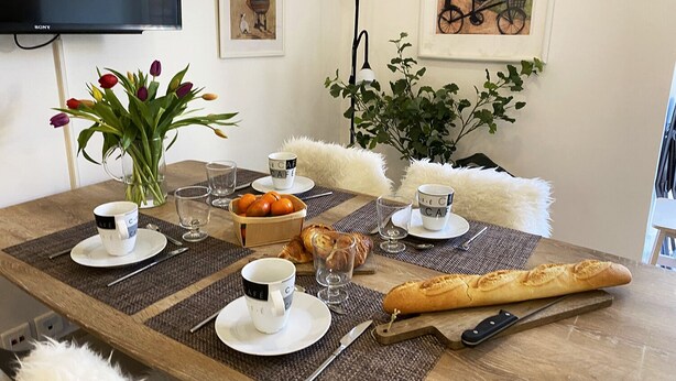 Une table à manger dressée avec des tasses à café, un vase de tulipes, un panier d’oranges, une baguette et un pain.