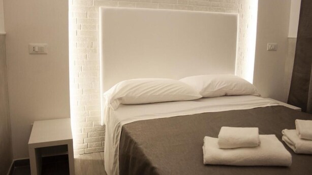 A neatly made bed with white linens and a brown bedspread, a small bedside table, and a white brick wall. - Rubino Guest House (Rome)