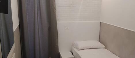 Una habitación pequeña y ordenada con una cama individual, una mesita, un control remoto y una lámpara fijada a la pared.