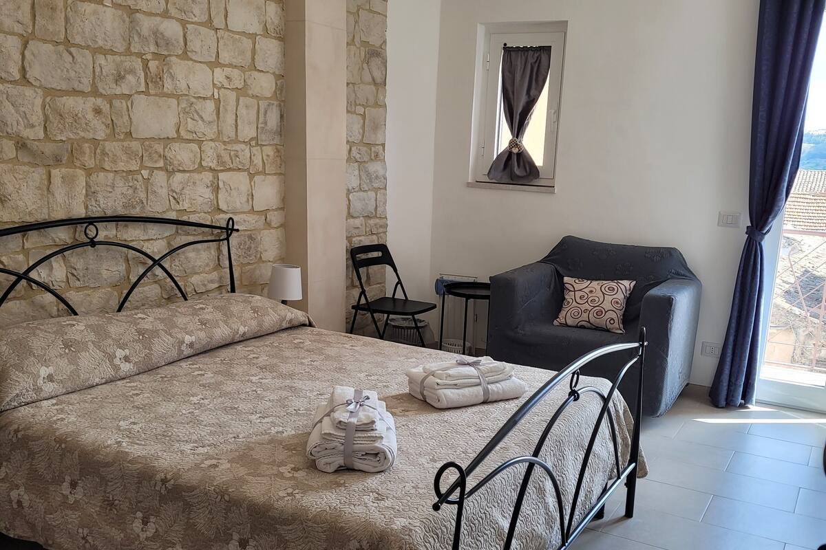 Un dormitorio con pared de piedra, cama con colcha floral, una silla, una mesita y un sofá.