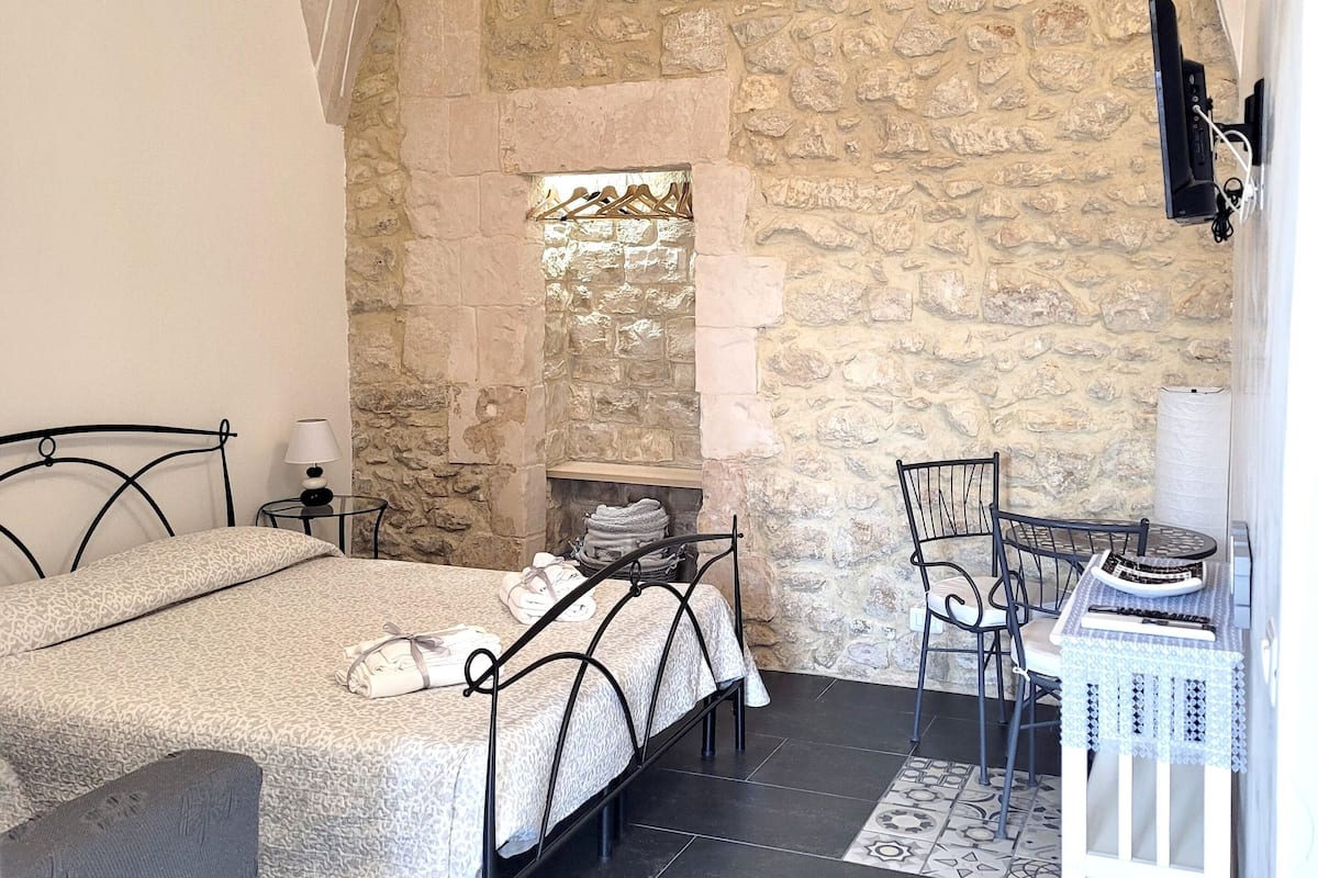 Un dormitorio con pared de piedra, una cama, una silla, una mesita y una chimenea.