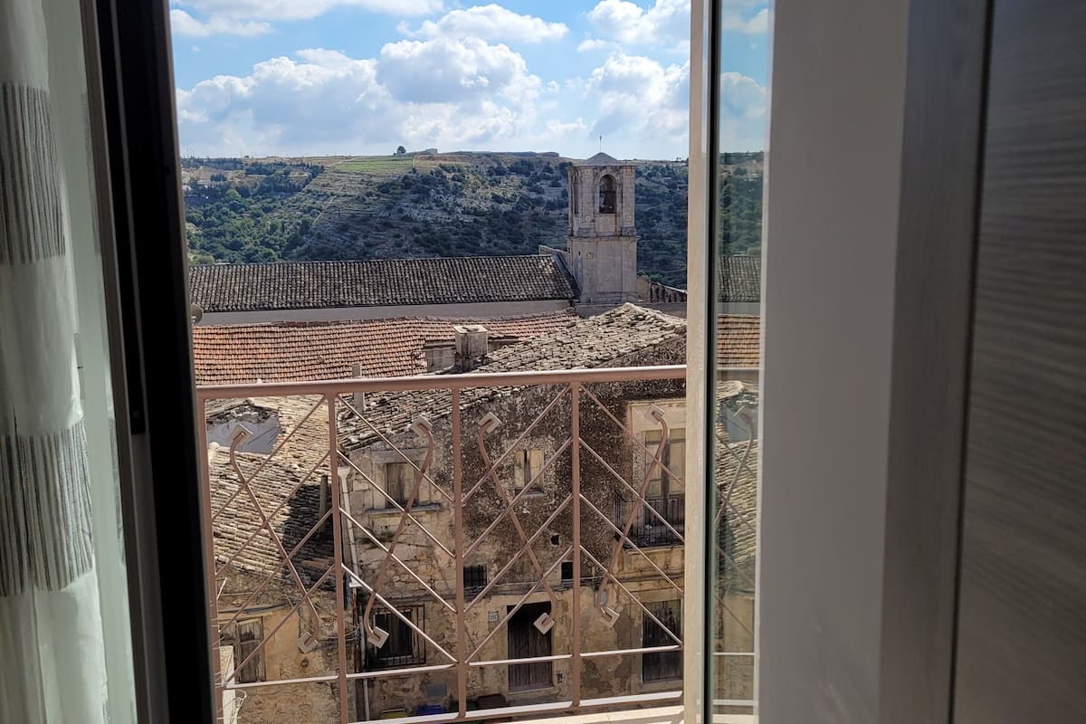 Vista desde un balcón con barandilla de metal, sobre un edificio histórico con una torre de campanas y tejados de tejas rojas.