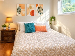 A bedroom with a patterned bedspread, a nightstand with a lamp, a potted plant, and wall art.