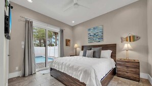 A bedroom with a large bed, a ceiling fan, a nightstand, a lamp, and a view of a swimming pool. - Iorana II | A Perfect Autumn Escape! | Game Room | Private Pool | Sleeps 21, Huge Private Master Suite! |  2 PM Check In!  |Gated Community with Amenities (Miramar Beach)