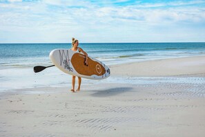 Une personne tenant une planche de paddle sur une plage de sable, avec l’océan en arrière-plan.