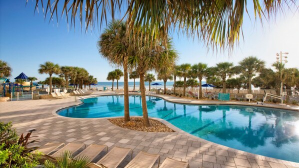 A swimming pool with a curved design, surrounded by palm trees and lounge chairs. - Good Times and Tan Lines! (Daytona Beach)