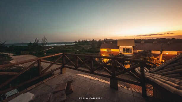 A sunset view from a balcony with a wooden railing, overlooking a beach and a building with warm lighting. - Pousada Pé na Areia (Garopaba)