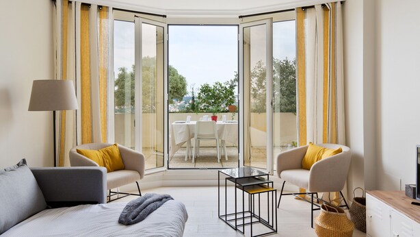 Una habitación de hotel moderna con un amplio ventanal corredero que da a una terraza exterior, una cama con cabecera gris, dos sillones con cojines amarillos, una mesita y una lámpara.