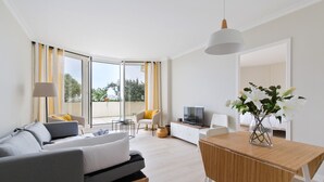 Un salón moderno con un sofá, una mesa de centro y un televisor. Se aprecia un balcón con cortinas amarillas y una silla.