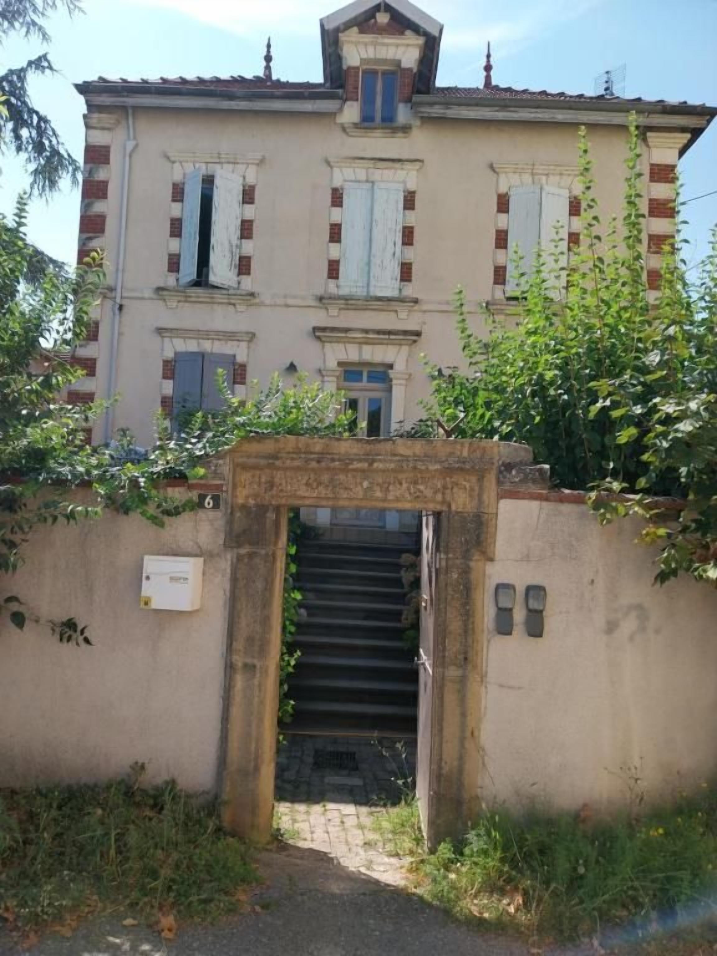 Un bâtiment à deux étages, doté d’un escalier central menant à une porte, le tout entouré de verdure.