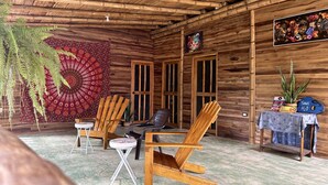 Una habitación rústica de madera con techo de paja, dos sillas de madera, una mesita y un tapiz en la pared.