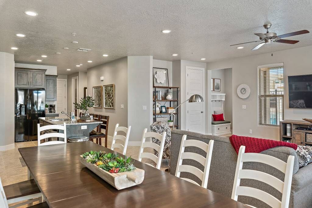 A spacious dining area with a wooden table and white chairs, a kitchen in the background, and a ceiling fan.