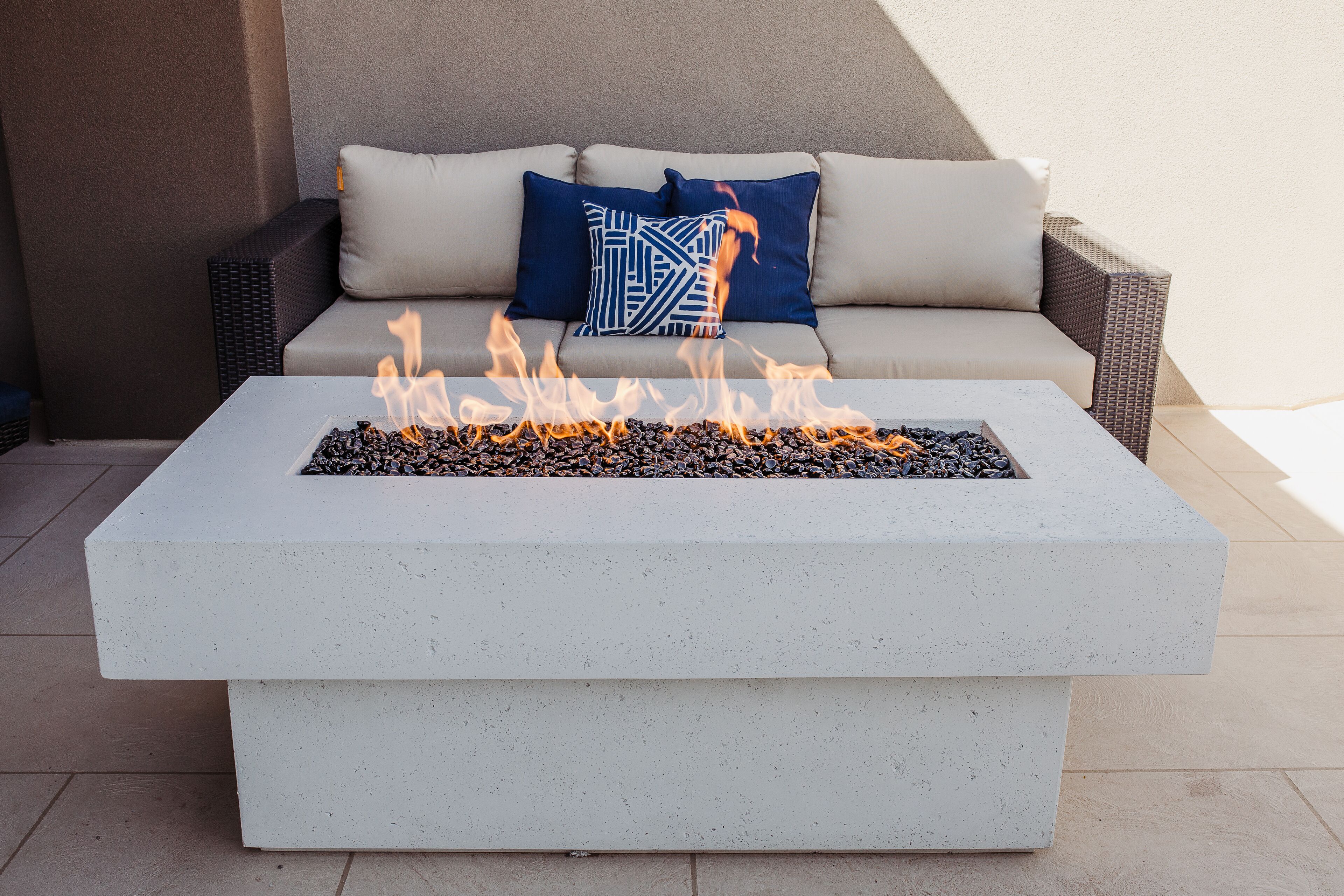 A concrete fire pit with flames, a beige sofa with blue pillows, and wicker furniture.