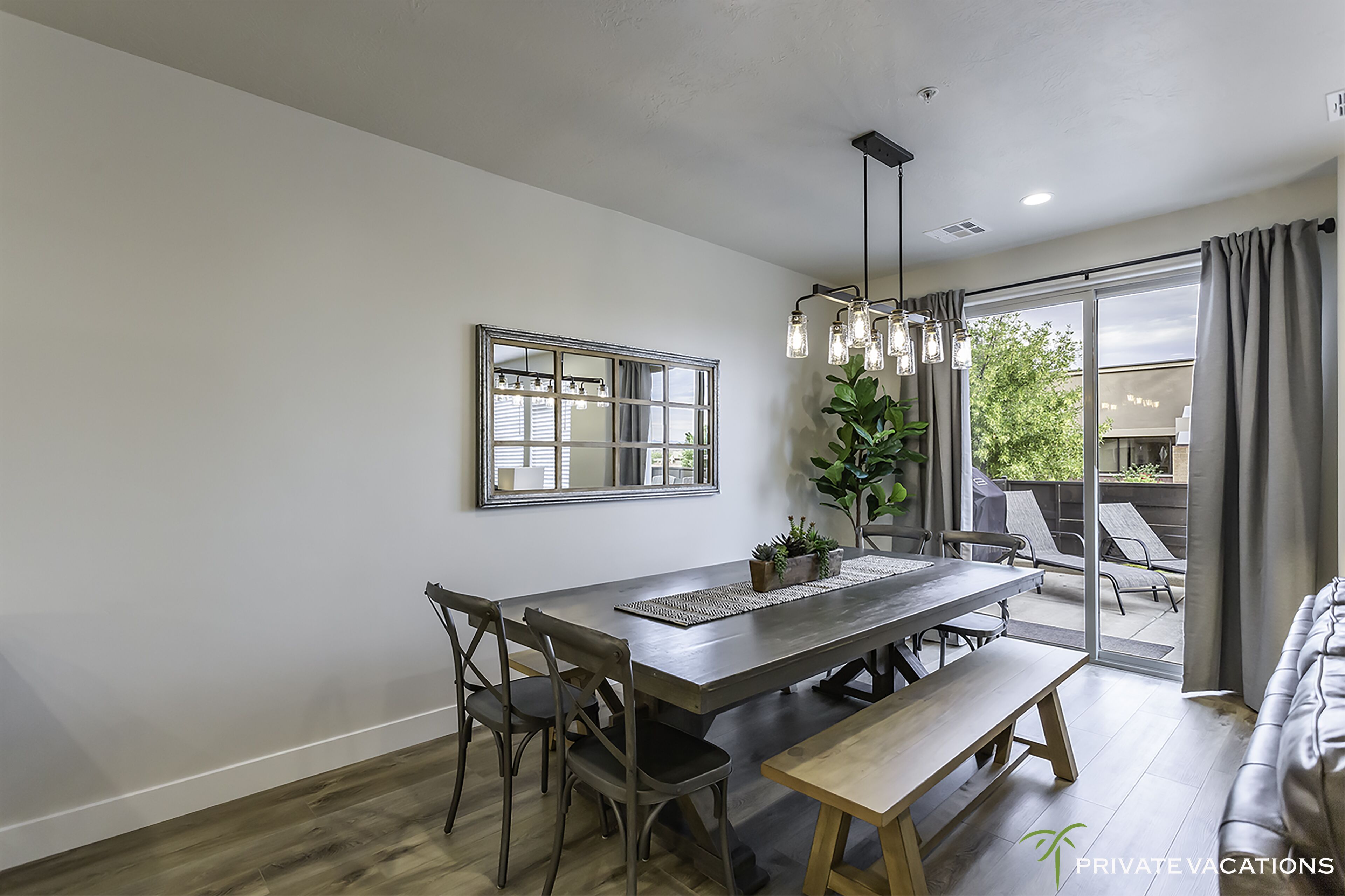 A modern dining area with a large table, chairs, and a bench. There is a large mirror on the wall, a hanging light fixture, and a view of the outdoors through a sliding glass door.
