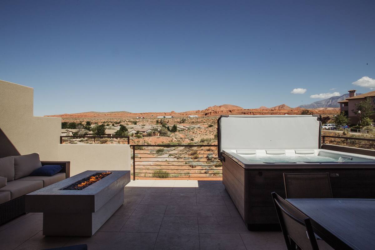 An outdoor patio with a fire feature, seating, and a hot tub overlooking a desert landscape.