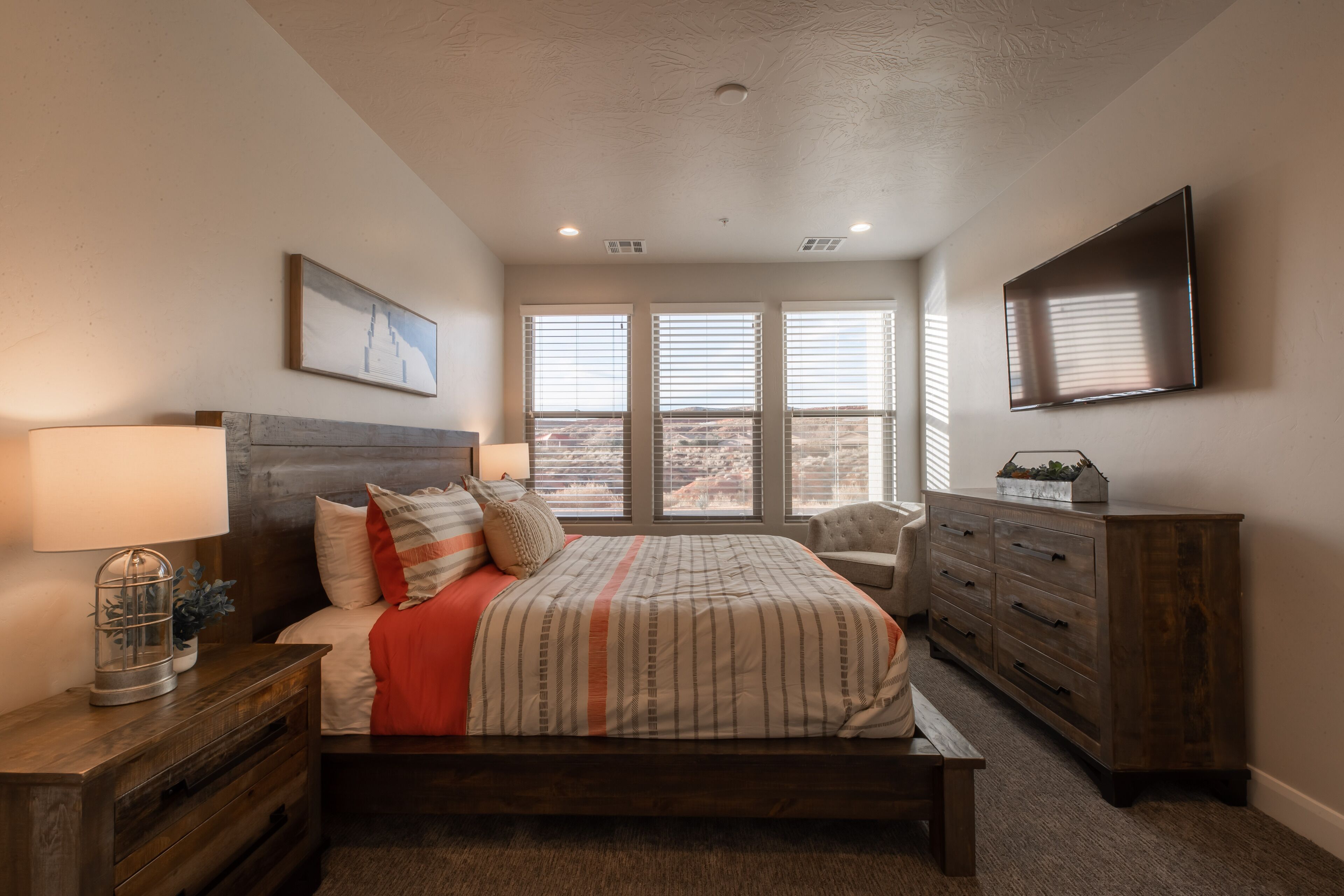 A bedroom with a bed, a nightstand, a dresser, a chair, and a TV on the wall.