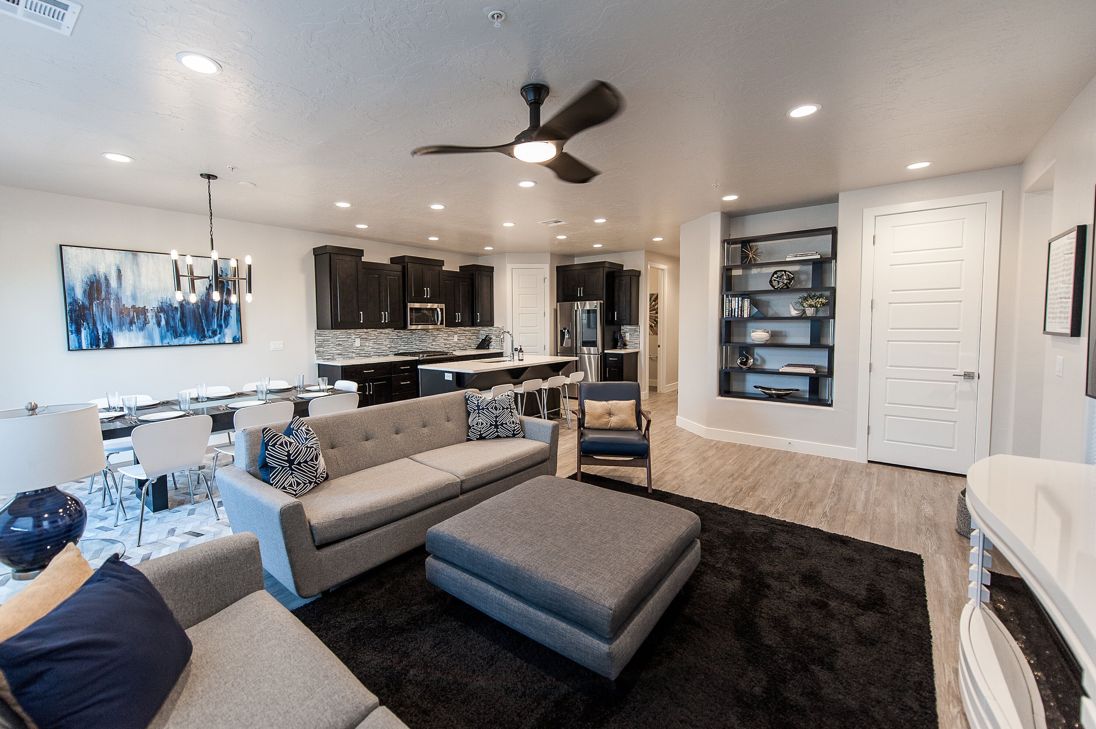 A modern living room with a sofa, ottoman, and chairs, a dining area with a table and chairs, a kitchen with dark cabinets and appliances, and a ceiling fan.
