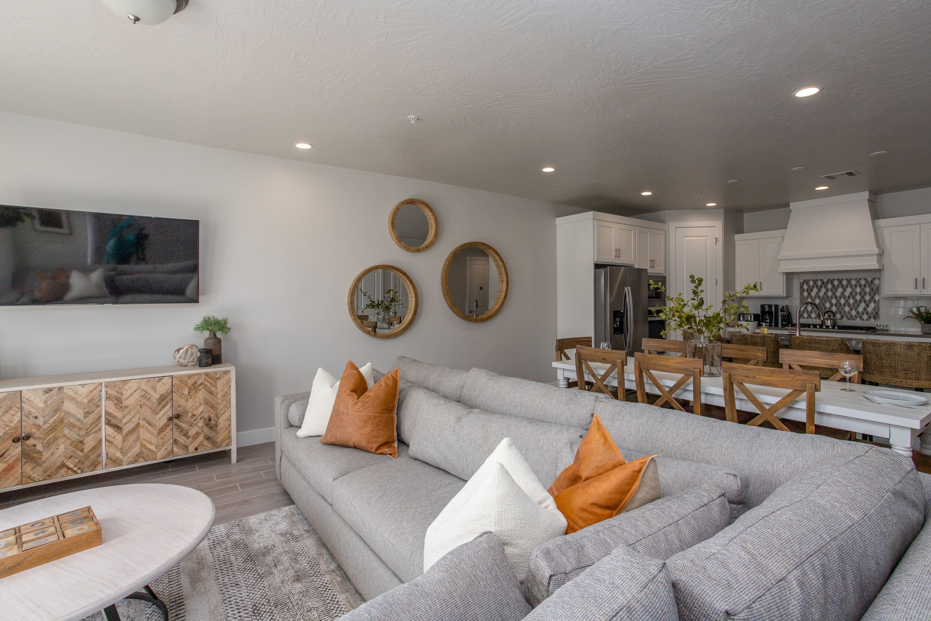 A modern living room with a grey sectional sofa, a herringbone-patterned cabinet, and a dining area with wooden chairs and a white table.