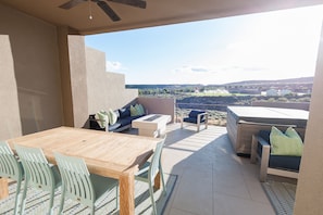 Eine Terrasse mit Holztisch, Stühlen, einer Couch und einem kleinen Tisch, alles auf einem gefliesten Boden mit Blick auf eine Landschaft.