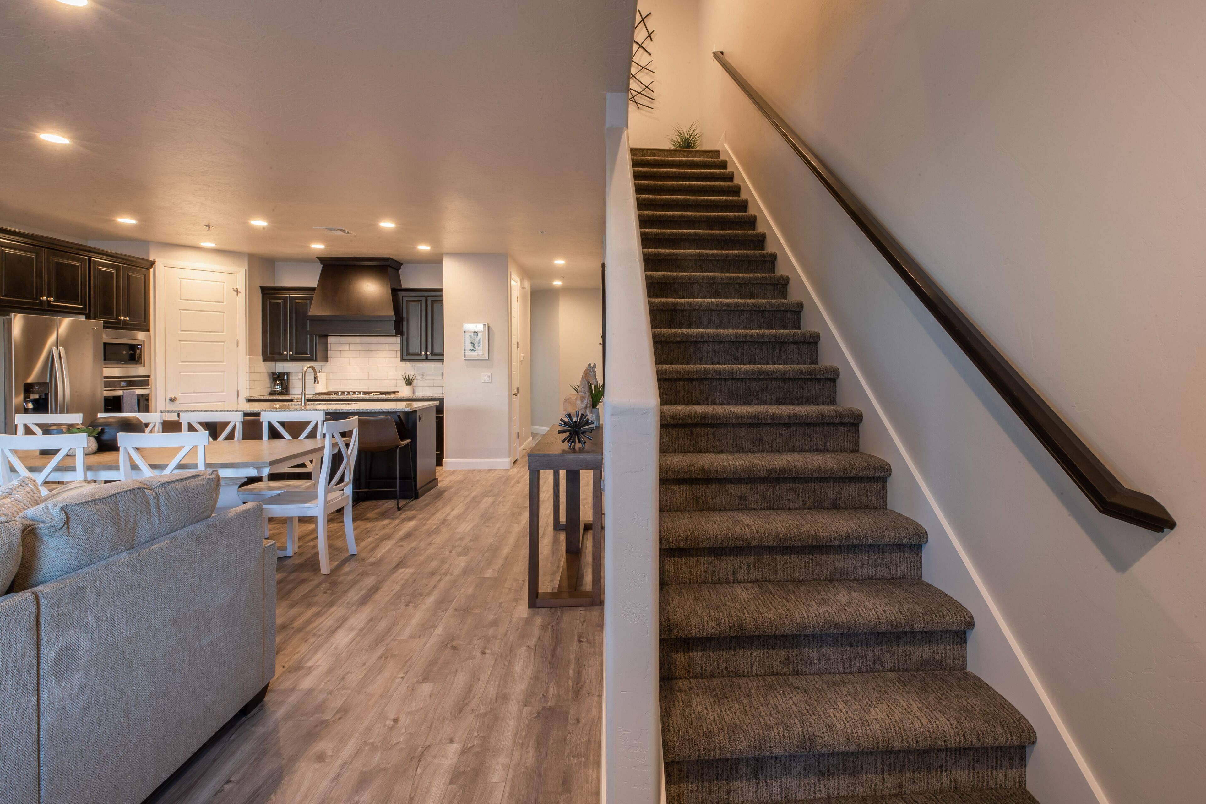 A modern kitchen with dark cabinets, a dining area with white furniture, and a carpeted staircase.