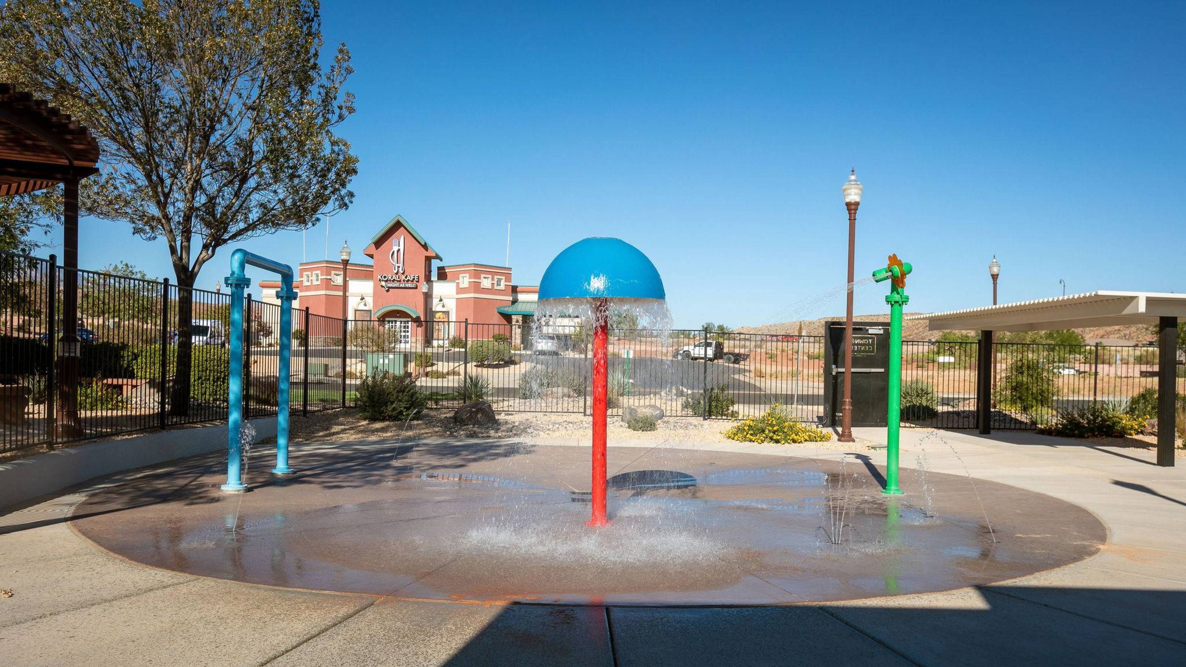 A water fountain with a blue dome and three colorful poles, surrounded by a paved area with a fence and a building in the background.