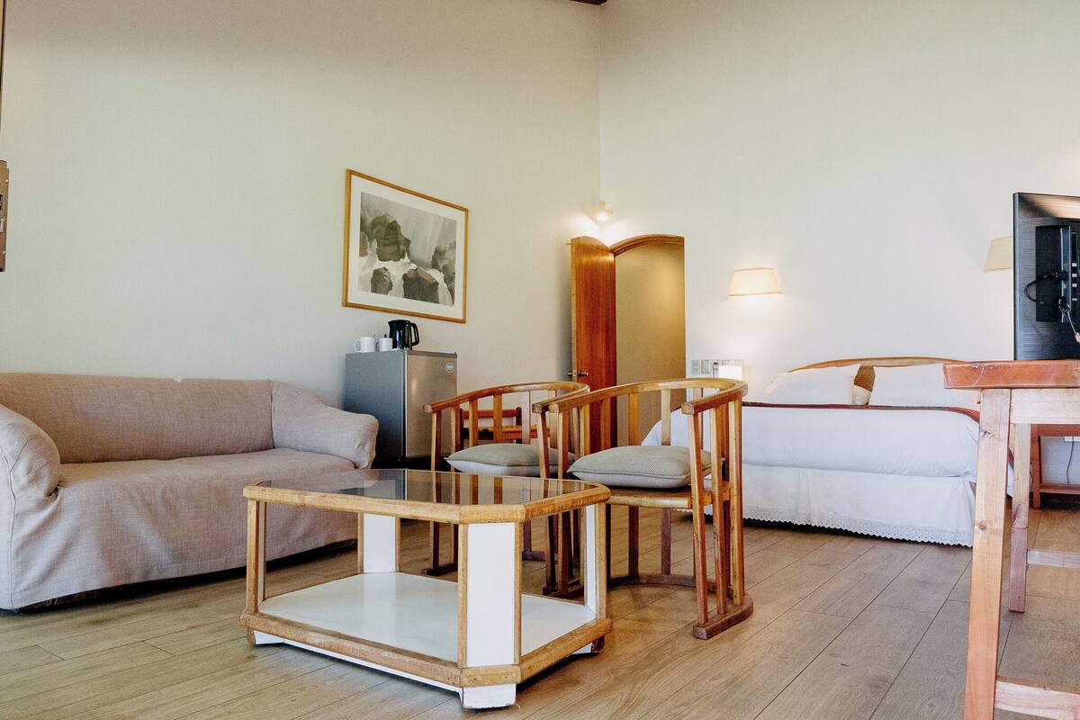 Una habitación acogedora con sofá, una mesa de centro de vidrio, sillas de madera, cama y espejo.