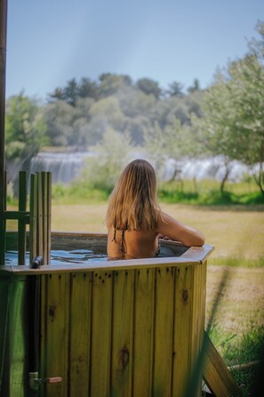 Eine Person entspannt in einer Holztub mit Blick auf einen Wasserfall und Bäume.