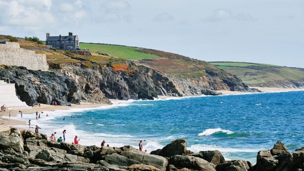 A coastal scene with a stone wall, beach, and a building on a hill. - Pipedream (Porthleven)