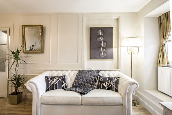 A living room with a white sofa, a framed mirror, a wall-mounted abstract art piece, a floor lamp, and a potted plant.