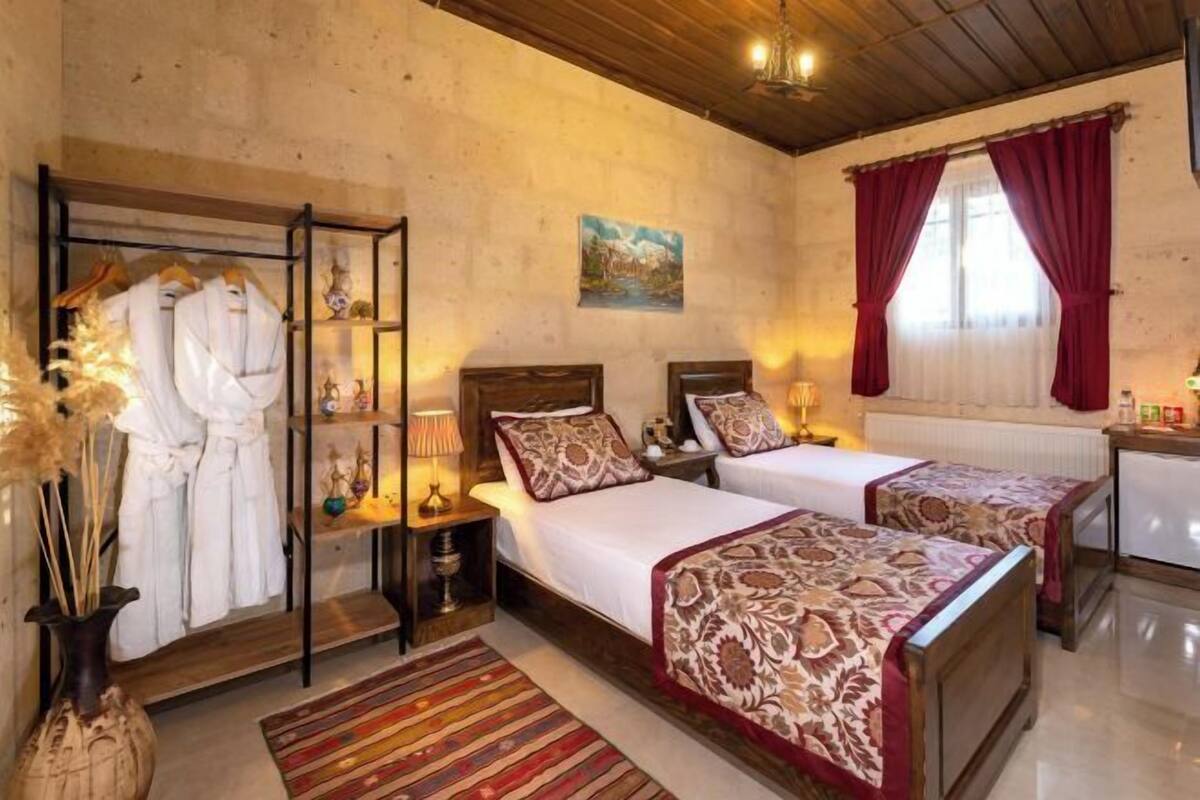 Huoneessa on kaksi sänkyä, puinen katto, seinällä taulu ja ikkuna, jossa on punaiset verhot.