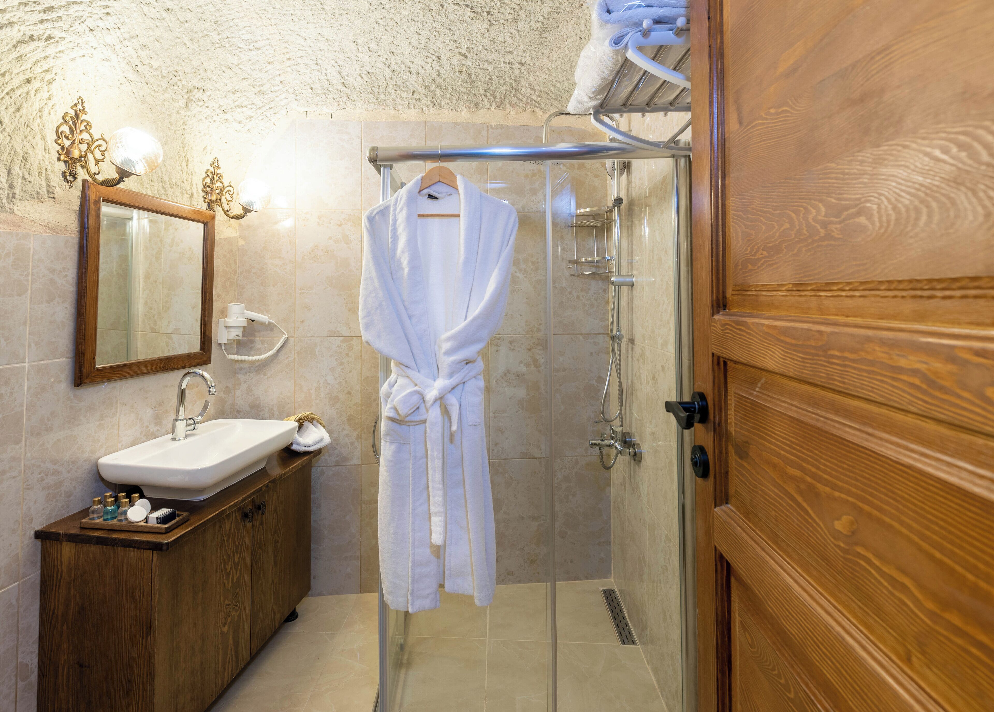 Un baño con tocador de madera, lavamanos, espejo, bata colgada en un perchero y cabina de ducha con mampara de vidrio.