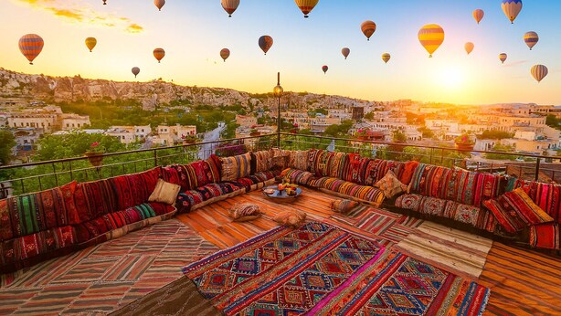 Une terrasse sur le toit, agrémentée de tapis colorés et d’un canapé, d’où l’on peut admirer un paysage urbain avec des montgolfières dans le ciel.