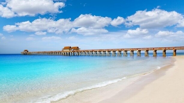 A beach with clear turquoise water, a sandy shore, and a wooden pier with buildings at the end. - Location and Lifestyle at its Finest! (Naples)