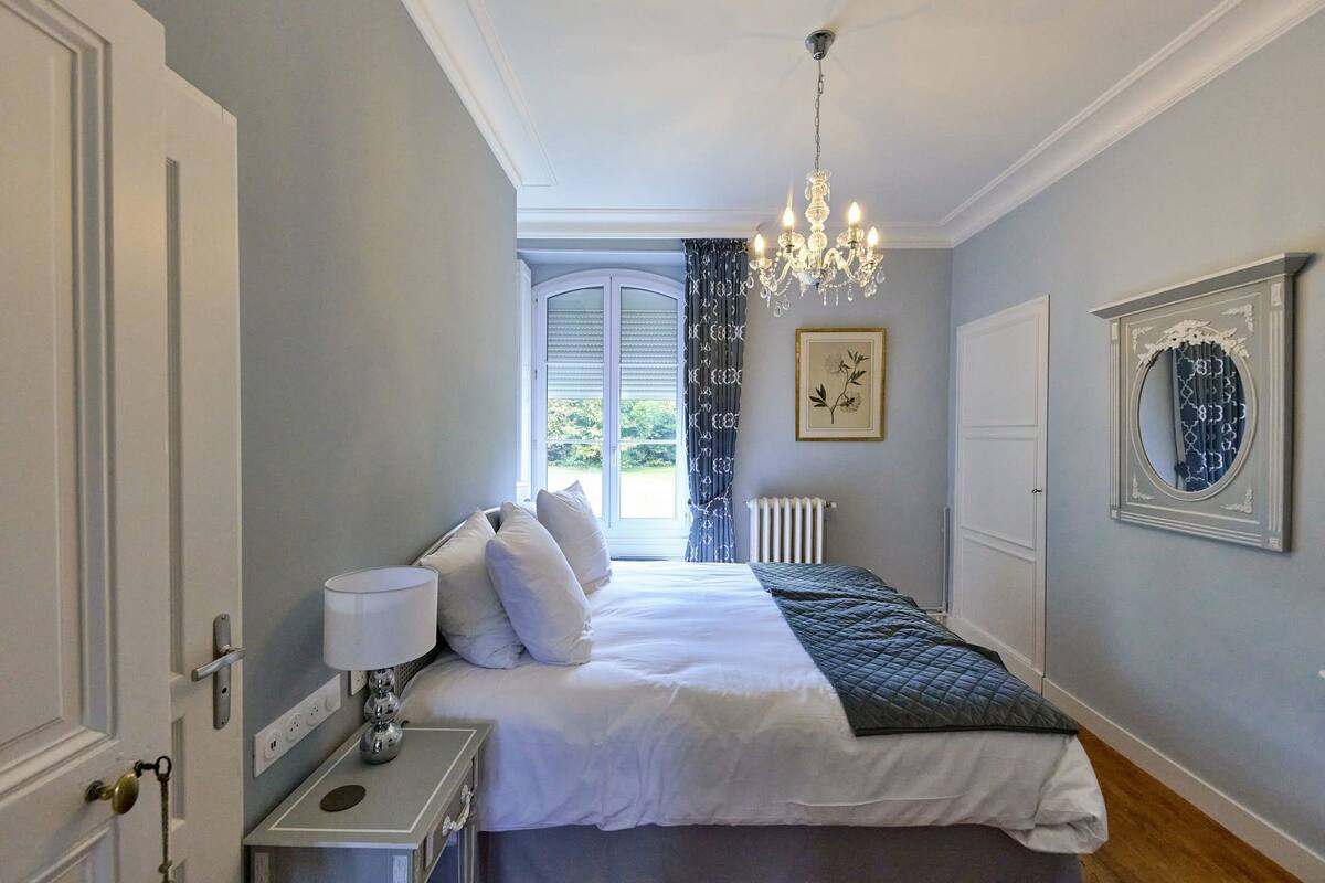 A bedroom with a large bed, a chandelier, a window with curtains, and a framed picture on the wall.