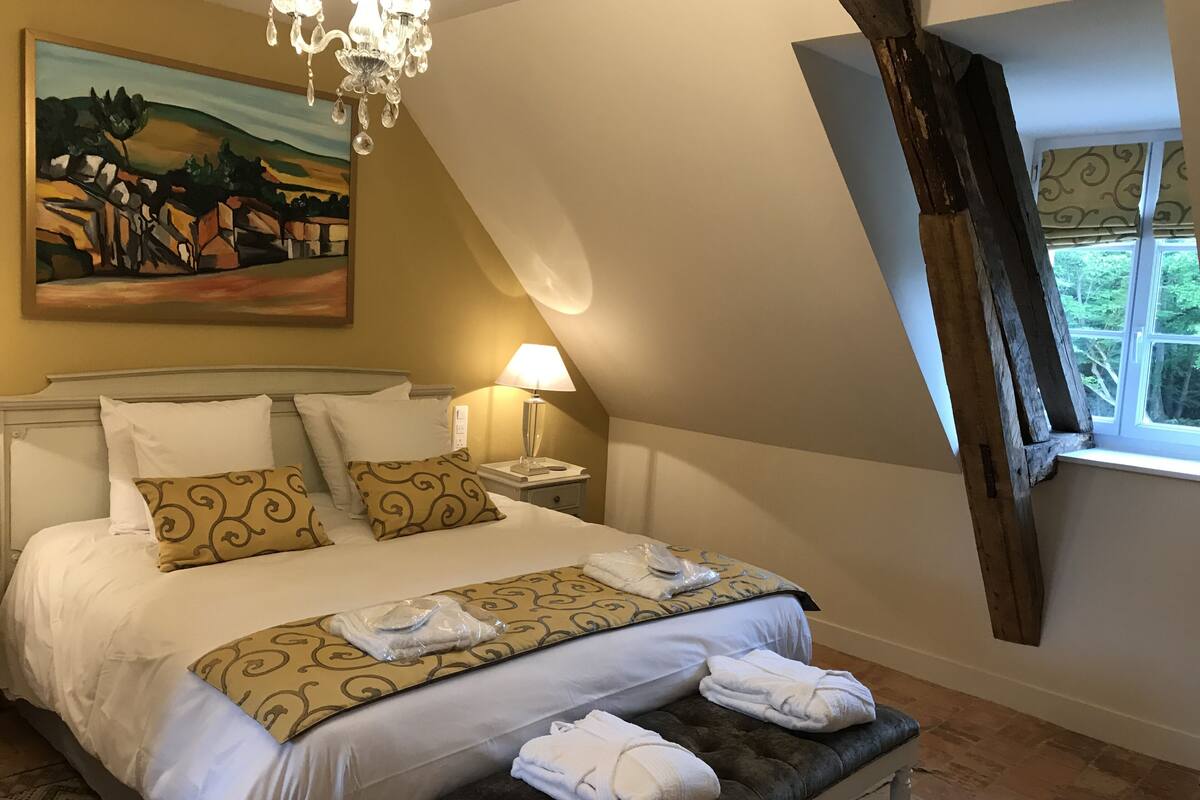 A neatly made bed with white and patterned pillows, a bedside lamp, and a bench with towels.