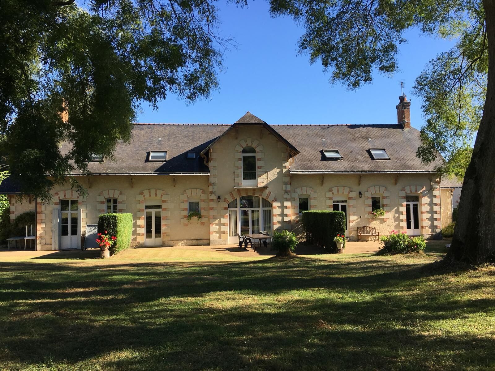 Ein großes, zweistöckiges Steinhaus mit einer symmetrischen Fassade, einem zentralen Eingang und einem kleinen Gartenbereich mit einer Bank und Topfpflanzen.