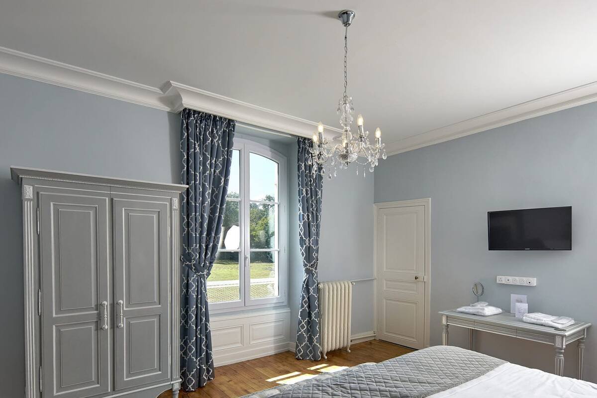 A bedroom with a large bed, a wardrobe, a desk with a flat-screen TV, and a window with curtains.