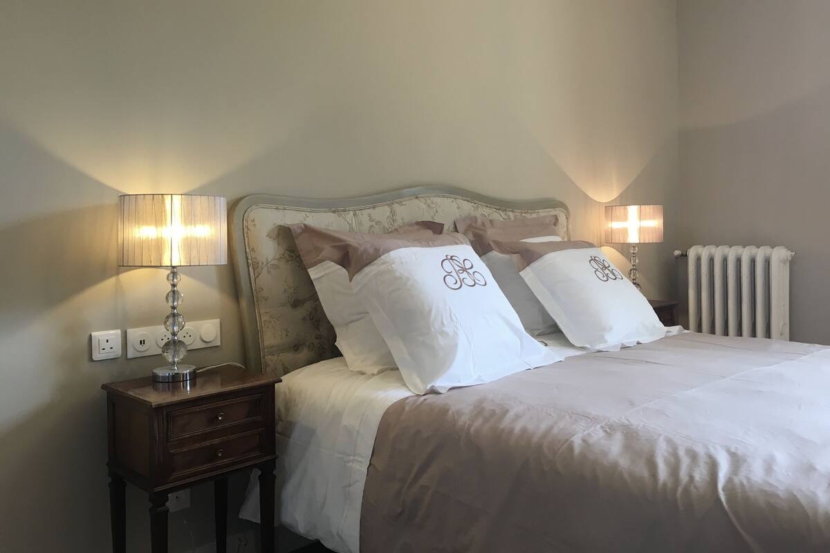 A neatly made bed with white linens and decorative pillows, a bedside table with a lamp, and a radiator on the wall.