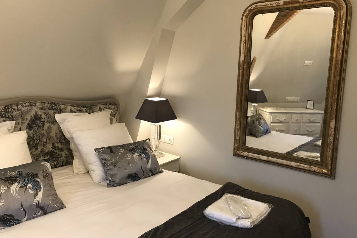 A hotel room with a bed, a large mirror, a nightstand with a lamp, and a dresser.