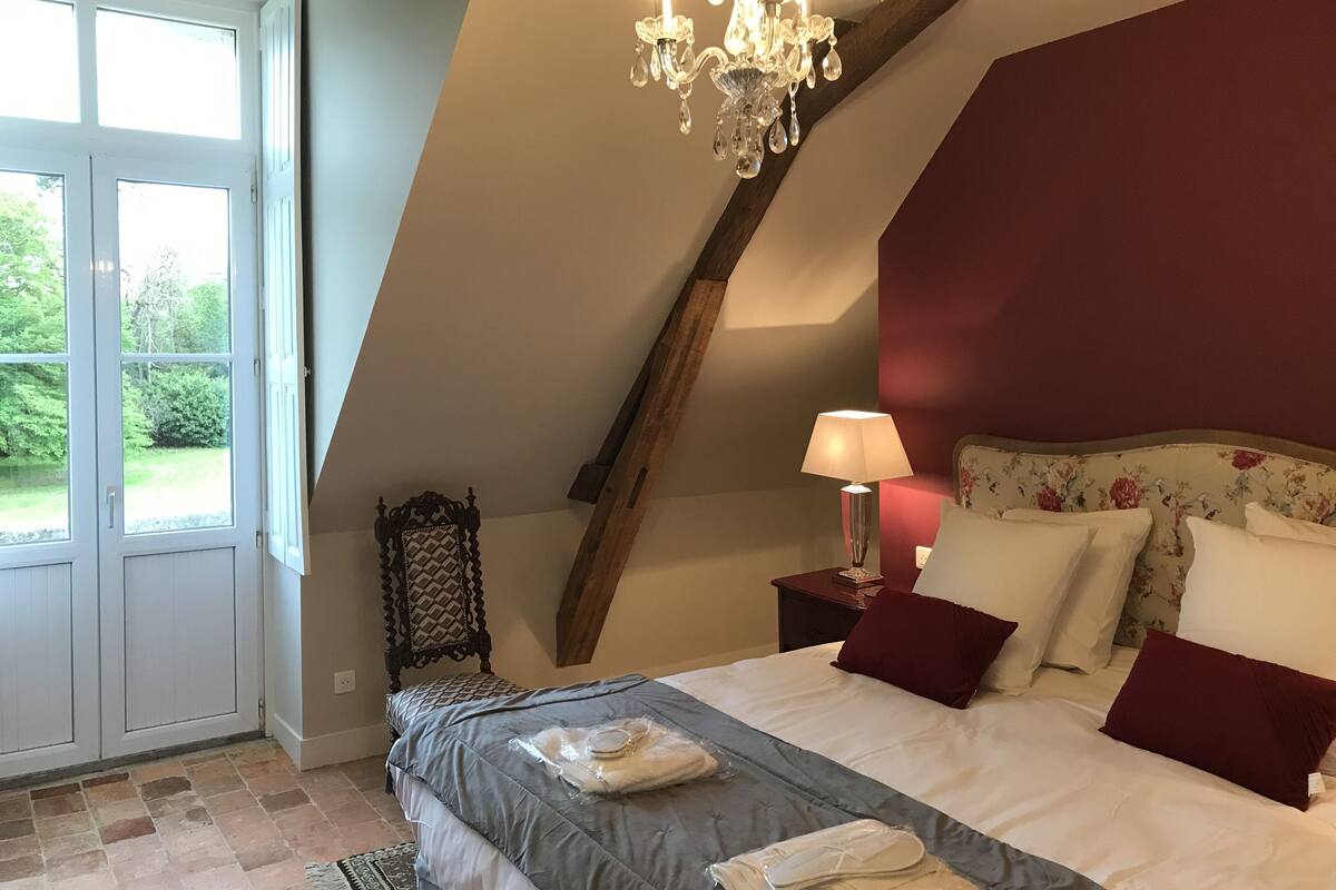 A bedroom with a sloped ceiling, a chandelier, a bed with pillows, a nightstand, and a chair.
