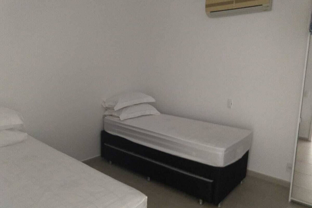 Ein kleines Zimmer mit einem Einzelbett, einer weißen Matratze, einem schwarzen Stauraum unter dem Bett und einer Wand-Klimaanlage.