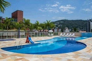 A swimming pool with lounge chairs and a slide, surrounded by palm trees and buildings.